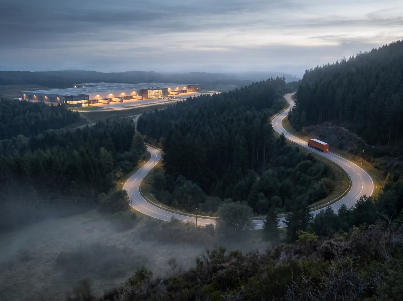 A lorry with an orange trailer still winding down a valley road toward a lit, empty dock at dusk, a week late