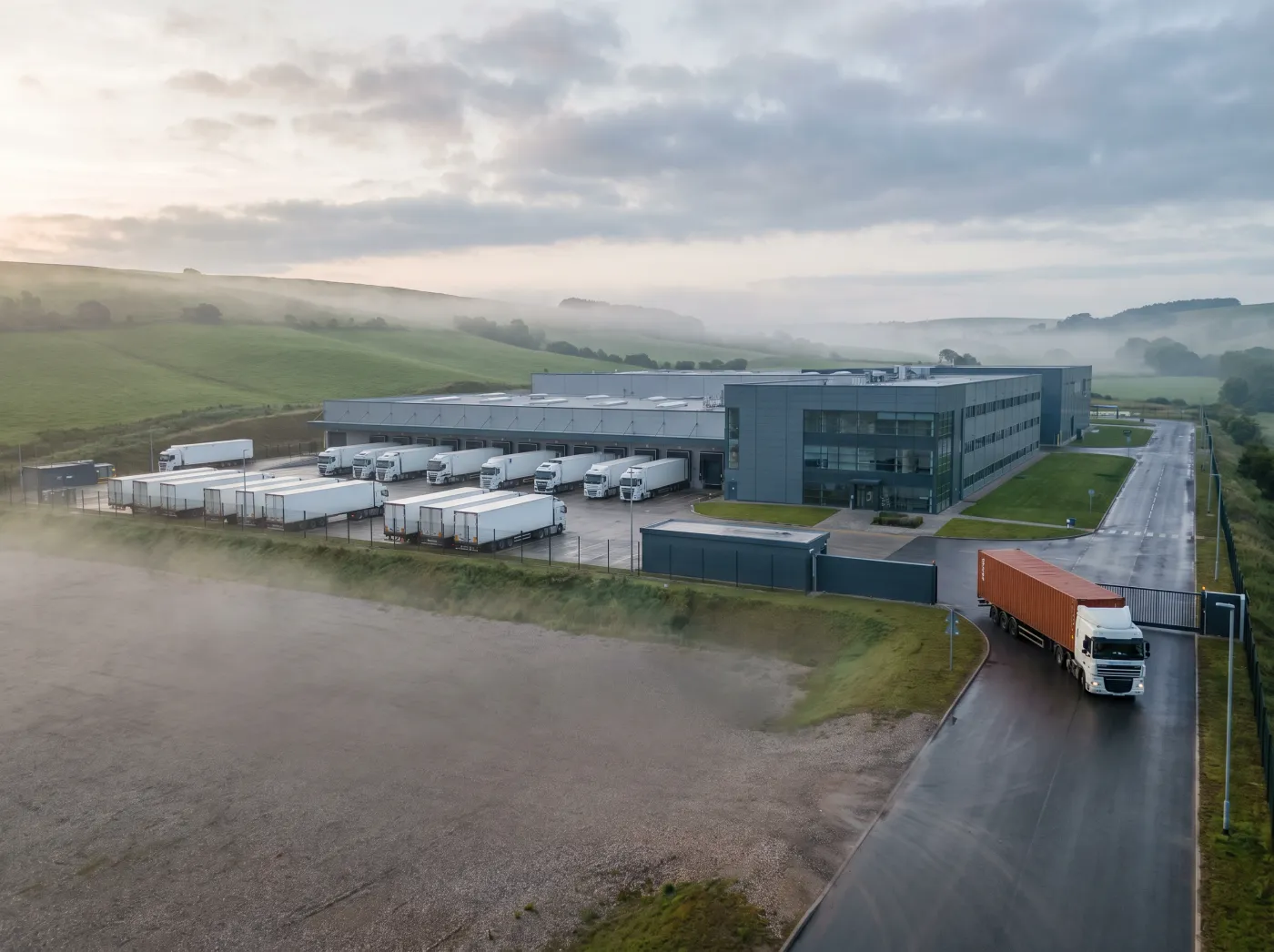 White lorries docked at a factory, one lorry with an orange trailer arriving late at the gate